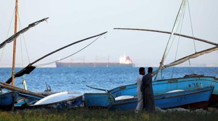 Fishermen walk in front of fishing boats as a container ship crosses the Red Sea before entering the Suez Canal, in Ismailia port city, northeast of Cairo, Egypt. 