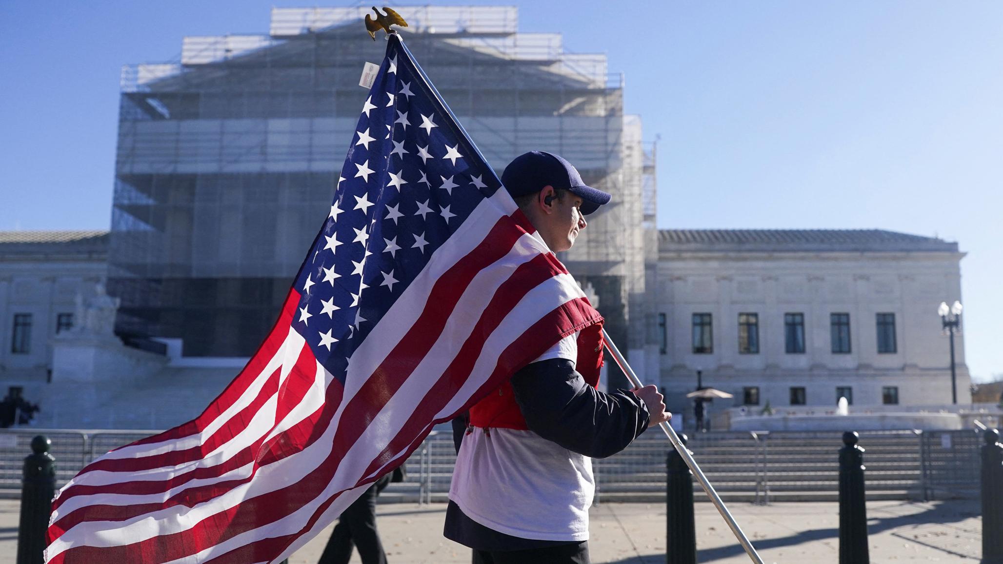 A man carries the US flag outside the US Supreme Court in Washington, DC, as its justices are set to hear oral arguments on President Trump's bid to preserve sweeping tariffs. Picture taken on November 5, 2025.