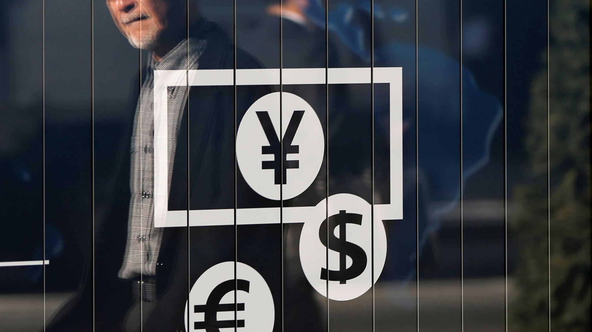 Passers-by are reflected on a signboard displaying currency signs outside a bank in Tokyo November 27, 2014. REUTERS/Issei Kato/File Photo